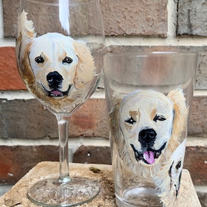 May include: Two glass cups with hand-painted portraits of golden retrievers. The wine glass features a close-up of a golden retriever's face, while the pint glass features a golden retriever looking to the side with another dog's head peeking out from behind.