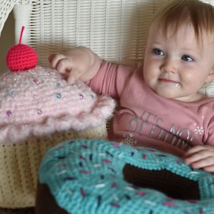 Op de afbeelding: Een gehaakte cupcake en donut met sprankels. De cupcake is roze met een rode kers bovenop. De donut is blauw met bruine glazuur. Een jong kind zit in een witte rieten stoel achter het gehaakte eten.