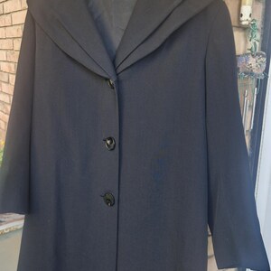 May include: A black coat with a wide collar and three buttons. The coat has three-quarter sleeves and is hanging on a wooden hanger. The coat is made of a smooth fabric.
