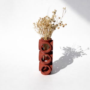 Peut inclure: Un vase rectangulaire de couleur terre cuite avec trois ouvertures circulaires, contenant des fleurs séchées. Le vase projette une ombre sur la surface blanche. Les fleurs séchées sont beiges et ajoutent un élément naturel au décor.