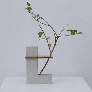 Peut inclure: Un vase en béton gris en forme de lettre L, doté d'un élément décoratif horizontal en bois avec une ouverture circulaire. Une fine branche dénudée avec de petites feuilles vertes est insérée dans le vase, s'étendant vers le haut et l'extérieur.