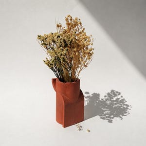 Puede incluir: Un jarrón geométrico de color terracota contiene flores secas en tonos beige y marrón. El jarrón tiene un diseño angular único y se encuentra sobre una superficie blanca. La sombra de las flores se proyecta sobre el fondo.