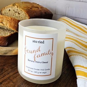 May include: A lit candle in a frosted glass jar with a label that reads "storied found family Banana Walnut Bread." Sliced banana bread sits on a wooden plate, with a loaf in the background. A yellow and white striped towel is on the right.