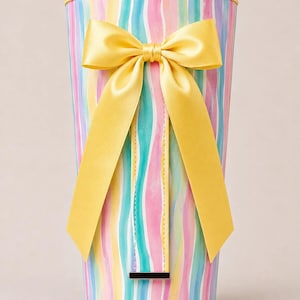 May include: A cylindrical container featuring a vertical wavy stripe pattern in pastel shades of pink, blue, and yellow. The top rim is trimmed in yellow, and a large, bright yellow satin ribbon bow is attached to the front center, above a small rectangular metal accent.