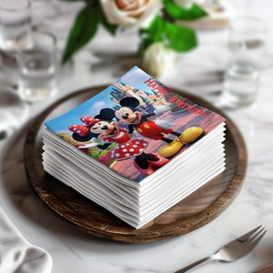 May include: A stack of white napkins with a colourful printed napkin on top. The printed napkin features Mickey Mouse and Minnie Mouse in front of a castle with the text "Happy Birthday".