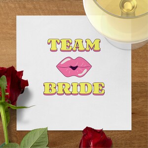 May include: White paper napkin with the text "TEAM BRIDE" in yellow letters with a pink lipstick kiss graphic in the center.