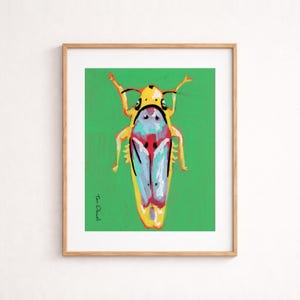 Op de afbeelding: Een ingelijste kunstdruk met een kleurrijke insectenillustratie. Het insect heeft een gele kop en lichaam, met blauwe en rode accenten op de vleugels, tegen een groene achtergrond. De handtekening van de kunstenaar, "Tom Duvall", is zichtbaar.