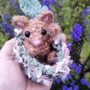 May include: A small brown crocheted animal with black bead eyes and a pink nose, nestled inside a textured green and pink crocheted basket. The animal has small ears and is accented with tiny silver sequins, while a green crocheted leaf sits behind it.