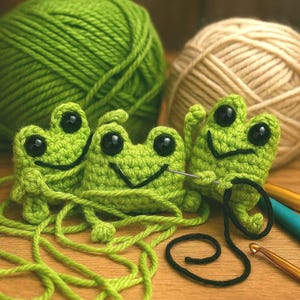 May include: Crocheted green frog toys with black eyes and smiling faces. The frogs are surrounded by green yarn, beige yarn, and crochet hooks, suggesting a handmade craft project. The frogs are connected by green yarn.