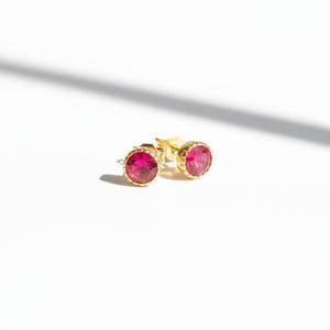 May include: A pair of gold stud earrings with round, vibrant pink gemstones. The earrings have a textured gold bezel setting, and are displayed on a white surface with a shadow.
