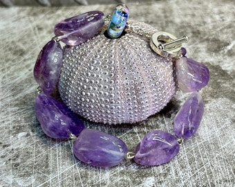 May include: A purple amethyst gemstone bracelet with a silver clasp. The bracelet is made of large, oval-shaped amethyst stones.