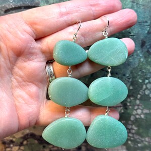 Peut inclure: Une paire de boucles d'oreilles pendantes avec trois perles de pierre vertes sur chaque boucle d'oreille. Les pierres sont de couleur vert clair et ont une finition lisse et polie. Les boucles d'oreilles sont en métal argenté.