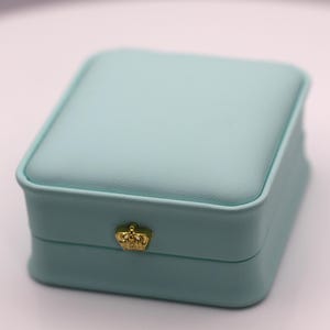 May include: A square, light blue jewelry box with a smooth, matte finish. The lid is slightly raised and padded. Centered on the front edge of the base is a small, metallic gold-colored crown emblem, serving as a decorative clasp detail.