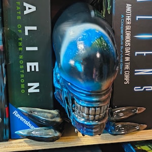 Op de afbeelding: Een blauwzwarte Alien-hoofd boekensteun met scherpe tanden en klauwen, die de Alien- en Aliens-speldozen ondersteunt. De gedetailleerde sculptuur is perfect voor fans van de Alien-franchise.