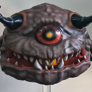 Op de afbeelding: Een bruin monster masker met een enkel rood oog, zwarte hoorns en een brede, tandige grijns. Het masker heeft een getextureerd oppervlak en is waarschijnlijk gemaakt van een duurzaam materiaal. Het ontwerp suggereert een wezen uit een fantasie- of horrorgenre.