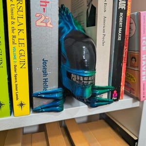 May include: A blue and green alien head bookend is placed between books on a white shelf. The bookend has a detailed design with sharp teeth and a glossy finish. Books include titles like "American Psycho" and "Catch-22".