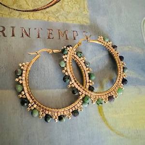 May include: Gold-tone hoop earrings with green and black gemstone accents. The hoops are embellished with small square gold beads and feature a hinged closure. The earrings are set against a background with the word "PRINTEMPS".