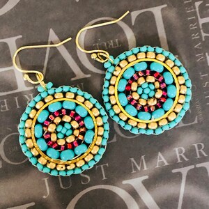 May include: A pair of circular beaded earrings with gold-coloured hooks. The earrings have a concentric design of turquoise, gold, and red beads. The outer ring is turquoise, followed by gold, and the centre is red. The earrings have a bohemian style.