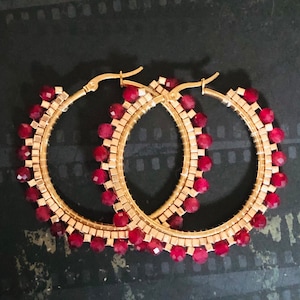 May include: A pair of gold hoop earrings with a red bead design. The earrings are made of metal and have a circular shape. The beads are small and round, and they are arranged in a pattern around the hoop.