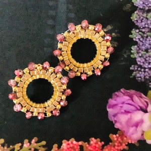 May include: A pair of gold and burgundy beaded earrings with a circular design. The earrings are made with small, square beads and round beads.