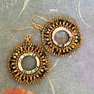 May include: A pair of gold hoop earrings with a circular design. The earrings are made of small, iridescent beads in shades of brown, gold, and green. The center of each earring features a round, iridescent shell.