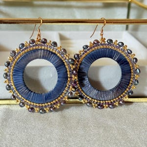 May include: A pair of handcrafted hoop earrings. The earrings feature a navy blue center, surrounded by a beaded border of gold, purple, and dark gray beads. The earrings have gold-colored ear wires.