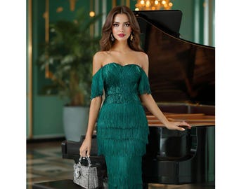 May include: A woman wearing a green off-the-shoulder dress with fringe detailing. The dress is fitted and has a sweetheart neckline. The woman is standing in front of a black grand piano.