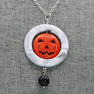 May include: A silver chain necklace with a pendant featuring a carved orange pumpkin face within a white and gray marbled circle. A black bead dangles below the pendant, creating a Halloween-themed accessory.
