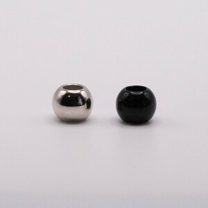 Puede incluir: Dos cuentas esféricas. Una es de color plateado y reflectante, la otra es negra y brillante. Ambas tienen un agujero en el centro. Estas cuentas probablemente se utilizan para la fabricación de joyas u otras manualidades.