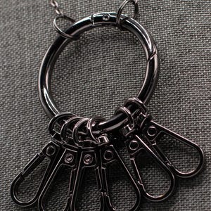Puede incluir: Un collar con llavero de color plomo con un gran anillo circular y seis pequeños clips tipo mosquetón. La cadena del collar también es de color plomo, sobre un fondo de tela gris texturizada.