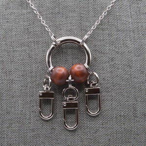 Puede incluir: Un collar de cadena plateada con un anillo plateado y tres llaveros plateados. El anillo tiene dos cuentas de madera marrón unidas a él.