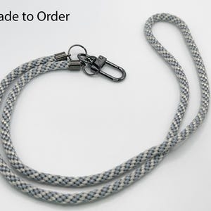 Può includere: Un cordino in corda grigia e bianca con moschettone e ferramenta in metallo nero. Il testo "Made to Order" si trova nell'angolo in alto a sinistra. Il cordino è realizzato in materiale di corda intrecciata.