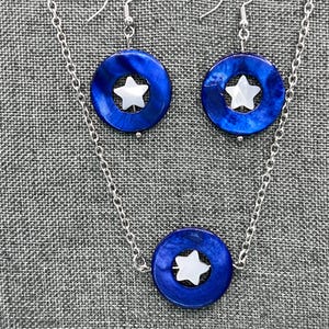 May include: A silver chain necklace and matching earrings set. The jewelry features a bright blue, circular pendant with a white star in the center. The earrings hang from silver hooks. The set is displayed on a gray textured surface.