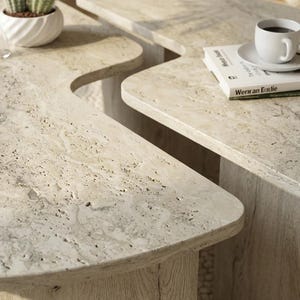 May include: Two beige travertine coffee tables with organic, curved edges and porous, textured surfaces. The tables feature light-colored wooden bases with visible grain patterns, supporting the heavy stone tops in a living room setting.