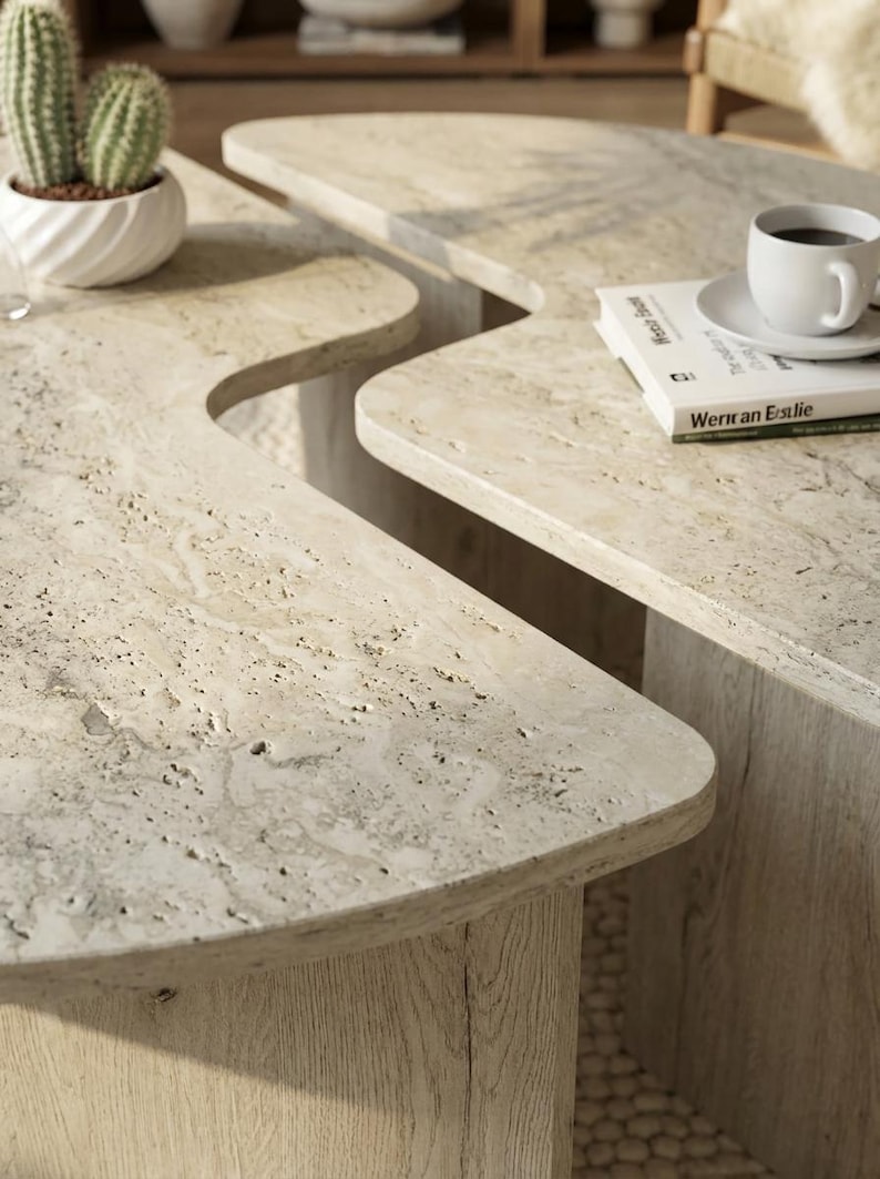 May include: Two beige travertine coffee tables with organic, curved edges and porous, textured surfaces. The tables feature light-colored wooden bases with visible grain patterns, supporting the heavy stone tops in a living room setting.