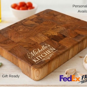 May include: A rectangular wooden butcher block cutting board with a personalized inscription that reads "Michelle's KITCHEN". The board is made of various wood pieces and has a natural wood grain pattern. A bulb of garlic and tomatoes are in the background.