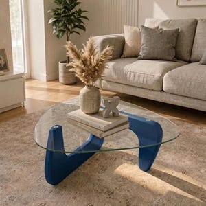 Peut inclure: Une table basse avec un plateau triangulaire en verre et une base en bois bleu. Sur la table se trouvent un vase beige texturé rempli d'herbe de la pampa séchée, deux livres empilés et un petit objet sculptural gris en forme de lettre X.
