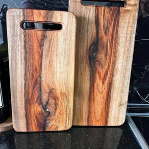 Puede incluir: Dos tablas de cortar rectangulares de madera con esquinas redondeadas y aberturas para asas integradas. Las tablas presentan un patr&oacute;n de veta de madera natural con vetas de color marr&oacute;n oscuro y nudos visibles, acabado pulido.