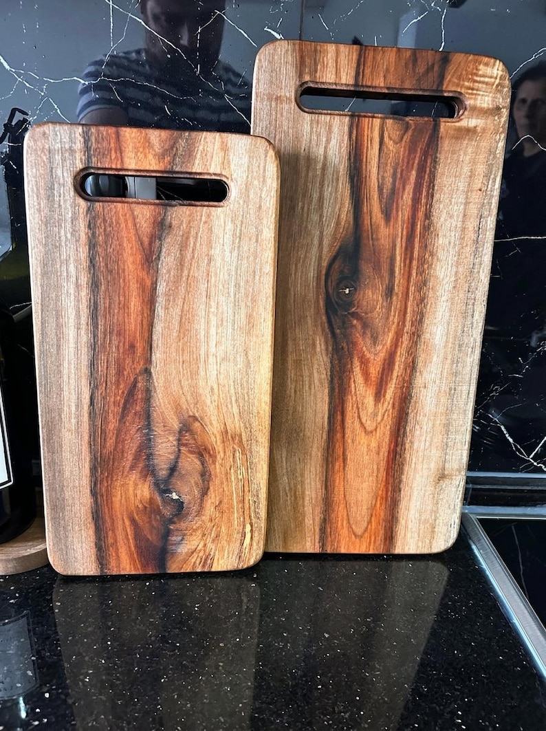 Puede incluir: Dos tablas de cortar rectangulares de madera con esquinas redondeadas y aberturas para asas integradas. Las tablas presentan un patr&oacute;n de veta de madera natural con vetas de color marr&oacute;n oscuro y nudos visibles, acabado pulido.