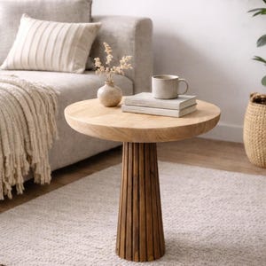 May include: A round, light-colored wooden side table with a fluted base. The table holds a mug, books, and a small vase with dried flowers. The table is 20 inches in diameter and 24 inches tall. The table is in a living room setting.