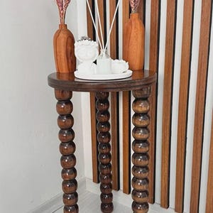 Op de afbeelding: Een ronde bijzettafel van donkerbruin hout met drie poten opgebouwd uit gestapelde houten bollen. Op het tafelblad staan twee hoge houten vazen met gedroogde bloemstengels, een wit keramieken beeldje en een glazen flesje met geurstokjes.
