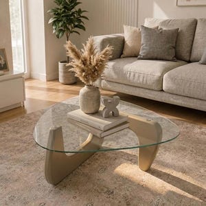 Peut inclure: Une table basse triangulaire en verre avec une base en bois clair. Sur le plateau se trouvent deux livres empilés, un vase en céramique beige texturé rempli d'herbes de la pampa séchées et un petit objet sculptural gris abstrait.
