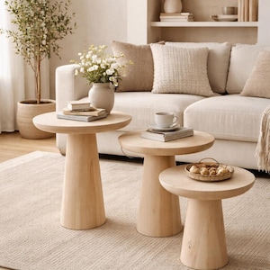 May include: A living room setting with three light-wood side tables. The tables have a round top and a pedestal base. The room also features a beige sofa, a woven pendant light, and a neutral-toned rug. The tables are in cm.