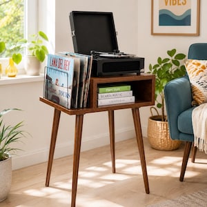Può includere: Un tavolino in legno marrone scuro con quattro gambe affusolate, dotato di un ripiano aperto contenente tre libri intitolati Plant Style, SCANDI RUSTIC e KINFOLK. Un giradischi portatile nero è posizionato sopra accanto a un supporto in metallo.