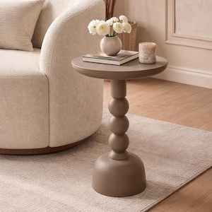 Può includere: Un tavolino color talpa con piano rotondo e base a sfere impilate. Ospita un piccolo vaso di fiori bianchi, un libro e una candela. Il tavolino è accanto a un divano color crema. Altezza: circa 60 cm.