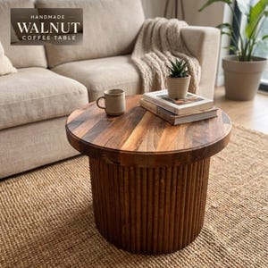 May include: A round walnut coffee table with a fluted, cylindrical base and a smooth, multi-toned wood grain top. The table holds a ceramic mug, two stacked books, and a small potted succulent. Text on the image reads: HANDMADE WALNUT COFFEE TABLE.