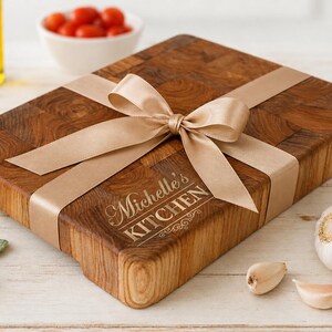 May include: A wooden cutting board with a butcher block design, wrapped with a tan ribbon and bow. The board is engraved with the text "Michelle's KITCHEN". Garlic cloves, a whole garlic bulb, and a bowl of cherry tomatoes are nearby.