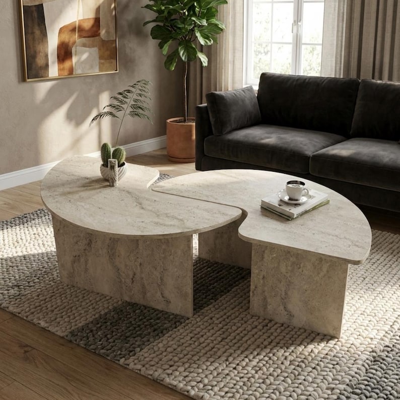 May include: A two-piece coffee table set featuring a beige travertine stone finish with a textured, porous surface. Each table has a curved, semi-circular top supported by a solid, matching stone base, creating a cohesive, organic shape.