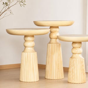 May include: Three light-colored wooden side tables of varying heights. Each table has a round top, a bulbous middle section, and a fluted base. The tables are arranged in a group, showcasing their different sizes and designs. The wood grain is visible.