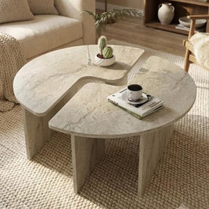 May include: A two-piece coffee table set featuring a light beige stone-like finish with natural veining. The tables are semi-circular in shape with vertical slab legs. A small potted cactus and a book with a white coffee cup sit on top.